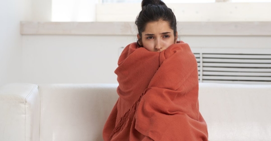 A person huddled up in an orange blanket near a heater.